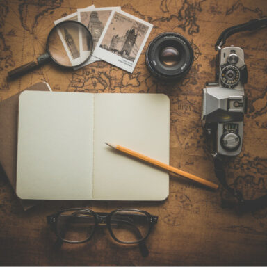 un cahier vierge avec un crayon a papier, un appareil reflex à coté ainsi qu'une loupe, des lunettes des photos en noir et blanc de londres sur une table représentant un planisphère